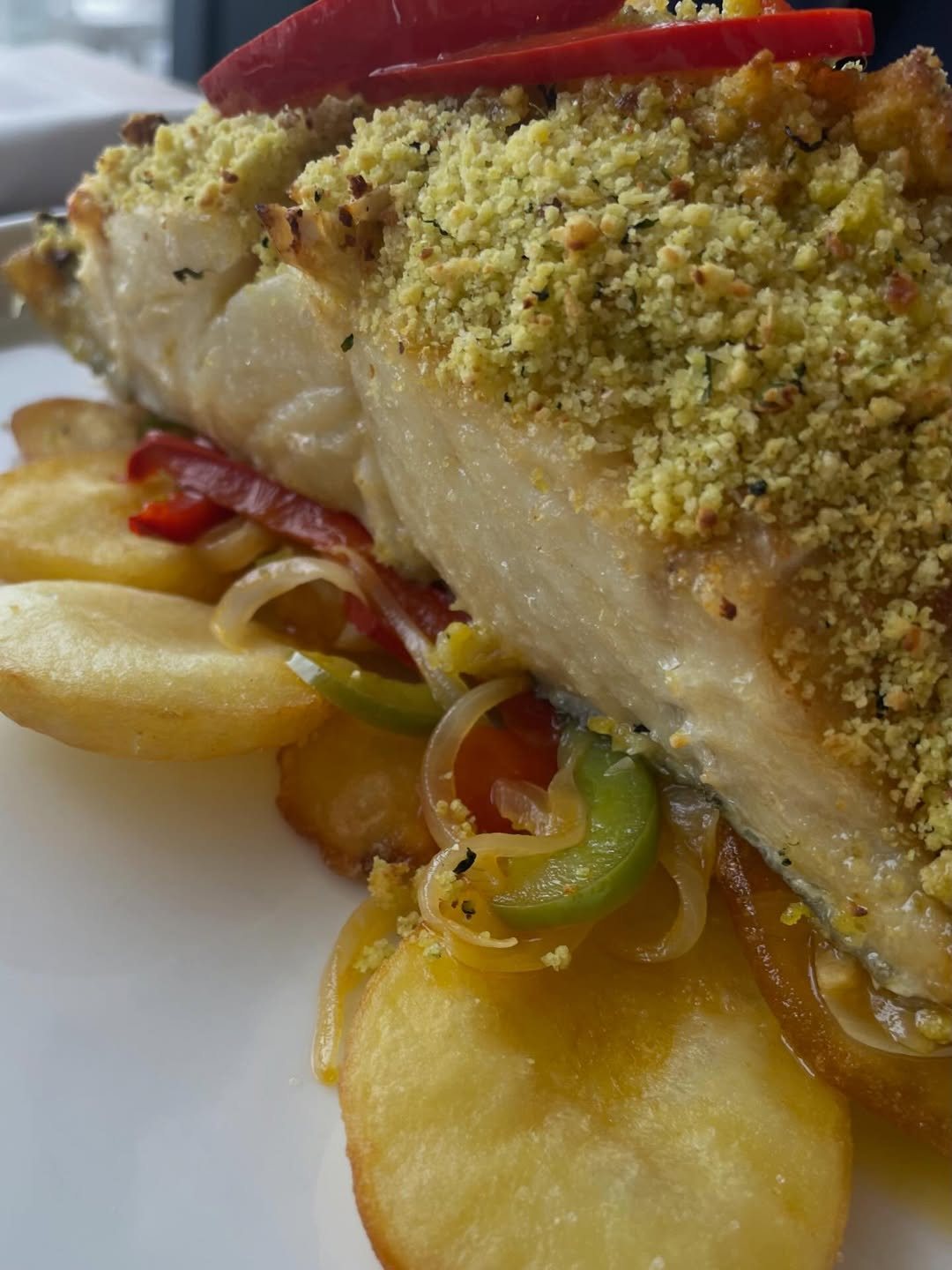 Posta de peixe com crosta dourada e esverdeada, sobre batata às rodelas, pimentos e cebola, num prato branco