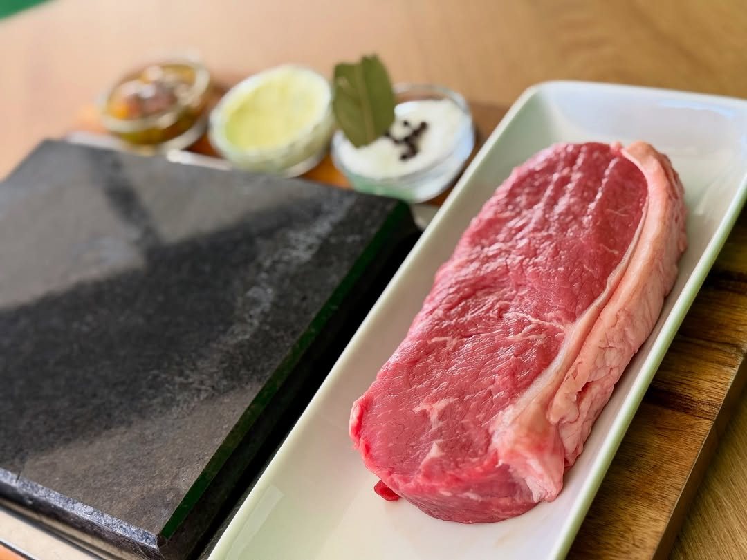 Bife cru sobre uma travessa branca, junto a uma pedra preta de grelhar e a pequenas taças com molhos