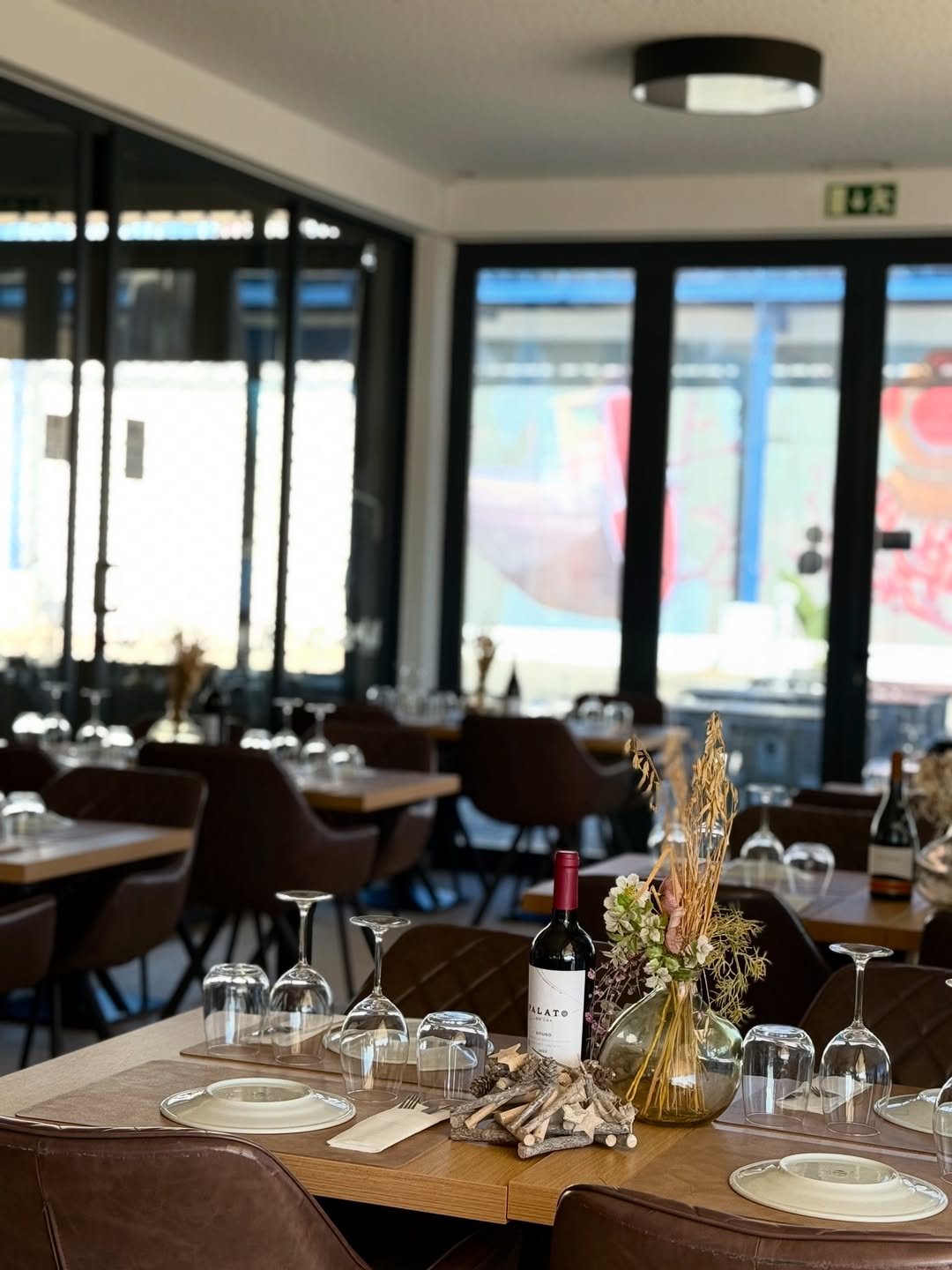 Sala do restaurante com mesas de madeira postas, copos, uma garrafa de vinho tinto e um jarro com flores secas, e grandes janelas ao fundo